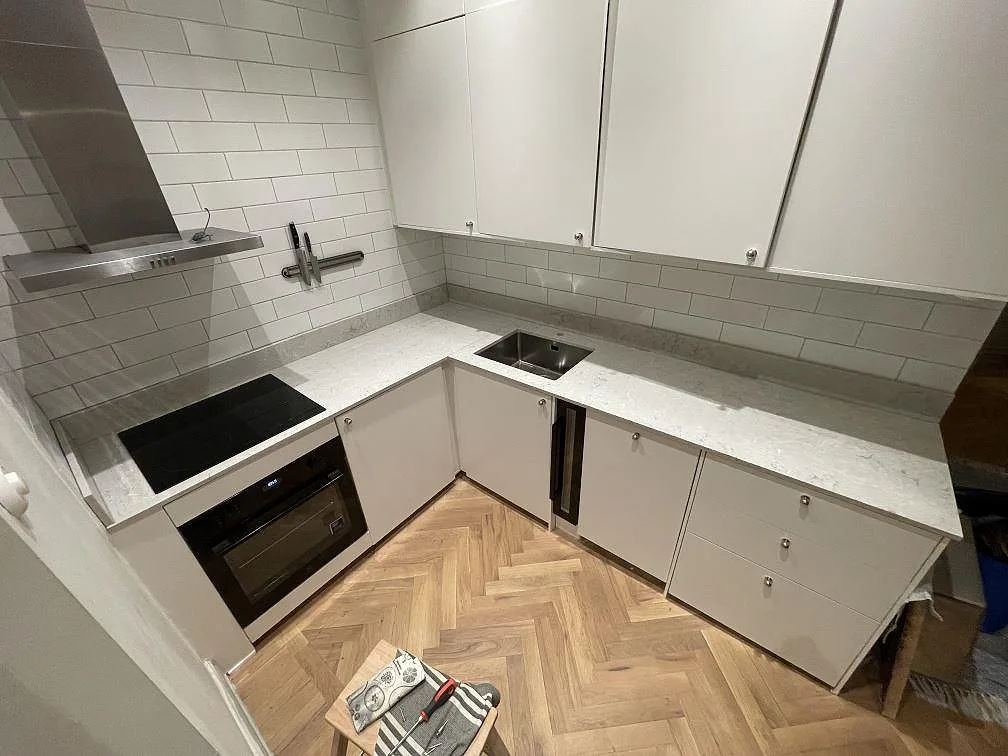 Köksbänkskiva i Caesarstone Bianco Drift, vit kvartskomposit med grå ådring, polerad, i ett L-format kök i Stockholm med underlimmad rostfri diskho och infälld induktionshäll mot vitt tegelkakel.