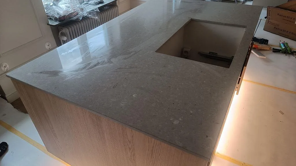 Stor köksö med polerad Caesarstone Clamshell i ljusgrå komposit, hälluttag och träinredning under montering i Malmö.