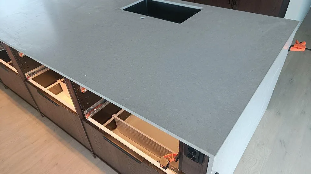 Topvy över köksö i matt Caesarstone Clamshell med förberedda urtag och öppna mörka skåpstommar i Nättraby.