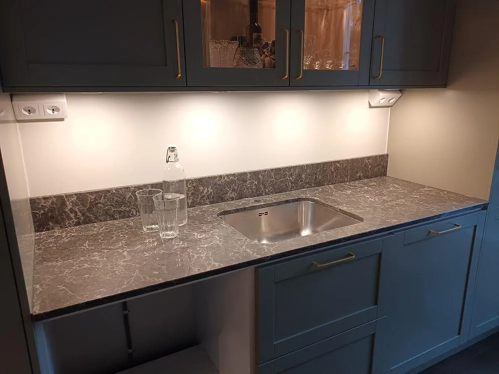 Köksbänkskiva i grå Caesarstone Coastal Grey, polerad kvartskomposit, med rostfri diskho och uppvikt stänkkant mot blågrå köksluckor i ett kök i Stockholm.