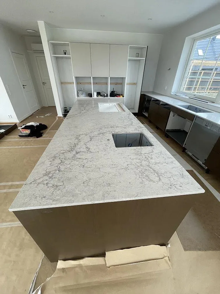 Fristående köksö i Caesarstone Moorland Fog, polerad ljusgrå kvartskomposit, sedd över hela köket med väggbänk och fönster i ett kök i Bunkeflostrand under montering.