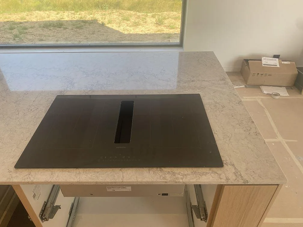 Köksbänkskiva i Caesarstone Moorland Fog, 20 mm polerad komposit, med svart induktionshäll och fönsterljus i ett kök i Farhult.