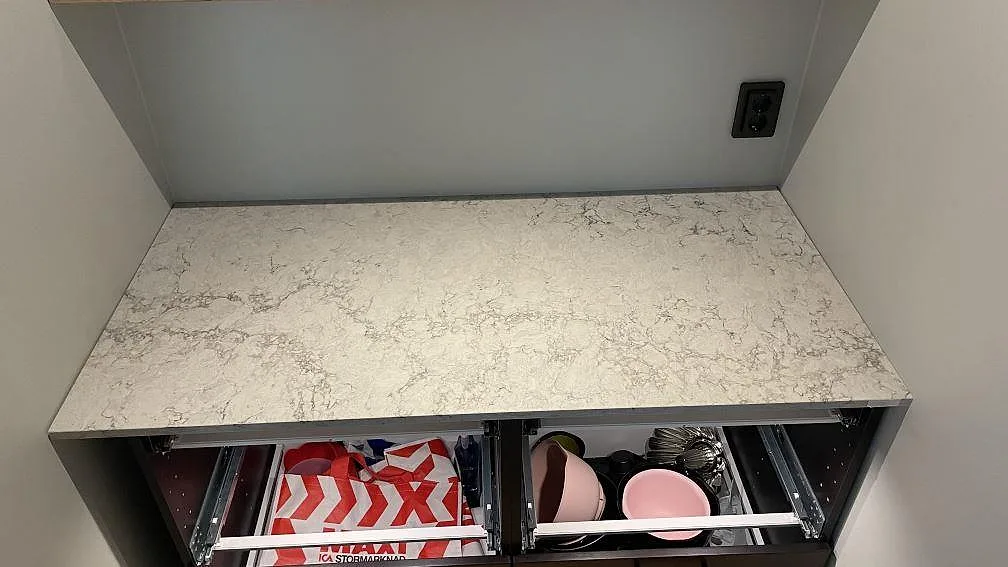 Rak bänkskiva i polerad komposit, Caesarstone Moorland Fog, monterad mellan väggar ovanför öppna stommar i Höllviken.