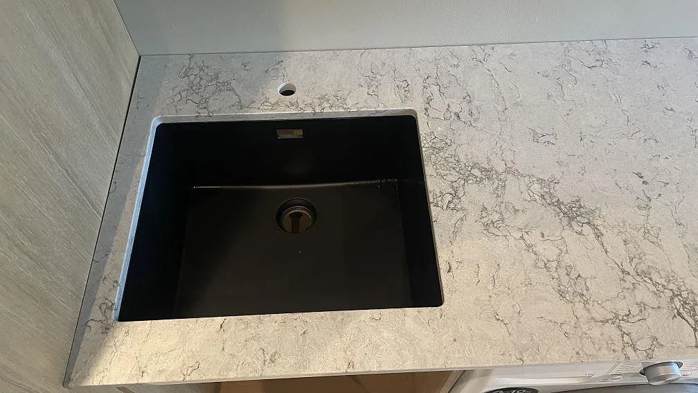 Närbild på svart diskho och blandarhål i polerad komposit, Caesarstone Moorland Fog, i en tvättstuga i Höllviken.