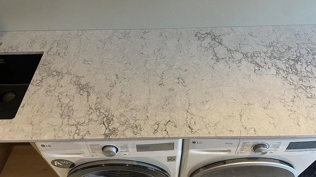 Detalj av ljus polerad komposit, Caesarstone Moorland Fog, med grå ådring över tvättmaskiner i Höllviken.