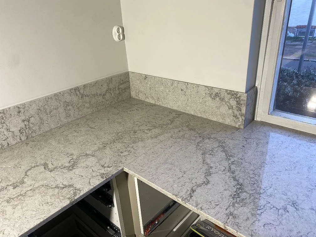 Polerad köksbänkskiva i Caesarstone Moorland Fog som vänder runt ett hörn med uppvikt stänkkant och fönster, i ett kök i Limhamn.
