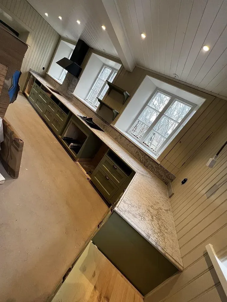 Översiktsbild av kök i Moheda med köksbänkskiva i polerad Caesarstone Moorland Fog, gröna luckor och vitt panneltak under montering.