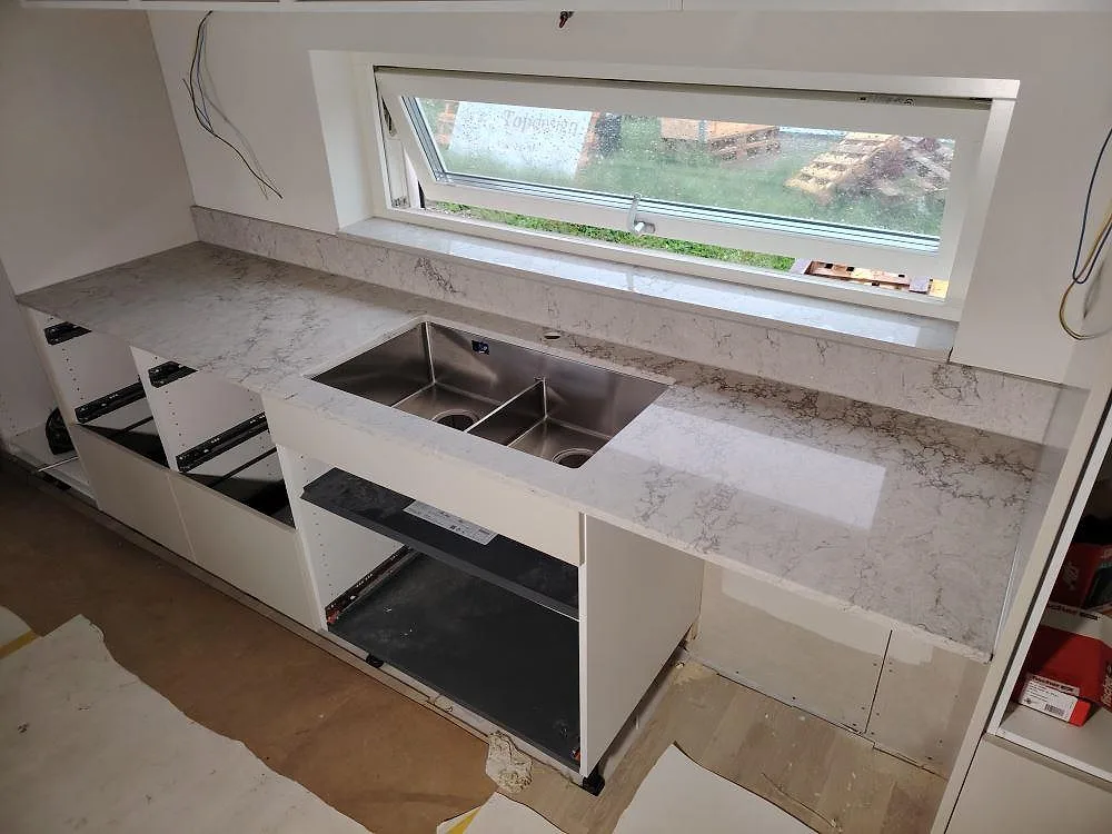 Väggbänk i Caesarstone Moorland Fog, polerad ljusgrå kvartskomposit, med låg stänkkant och rostfri dubbelho under fönster i Örebro.