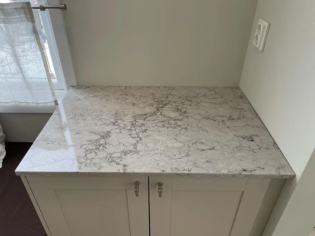 Köksbänkskiva i polerad Caesarstone Moorland Fog vid ett fönster, monterad på vit skåpstomme i ett kök i Saltsjö-Boo.