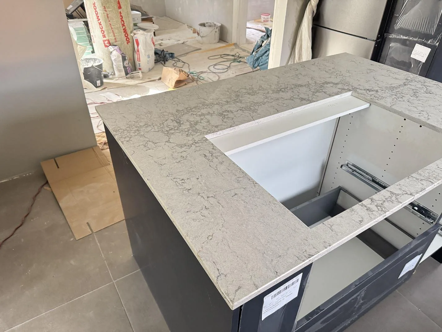 Köksö i polerad Caesarstone Moorland Fog, ljus kvartskomposit med grå ådring, med hällurtag i ett kök under montering i Svedala.