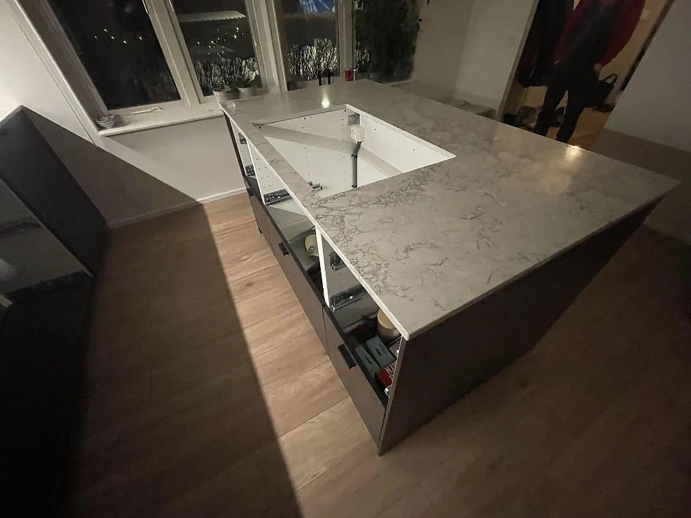 Köksö i polerad Caesarstone Moorland Fog med urtag för induktionshäll och öppna lådstommar, fotograferad under montering i ett kök i Tyresö.
