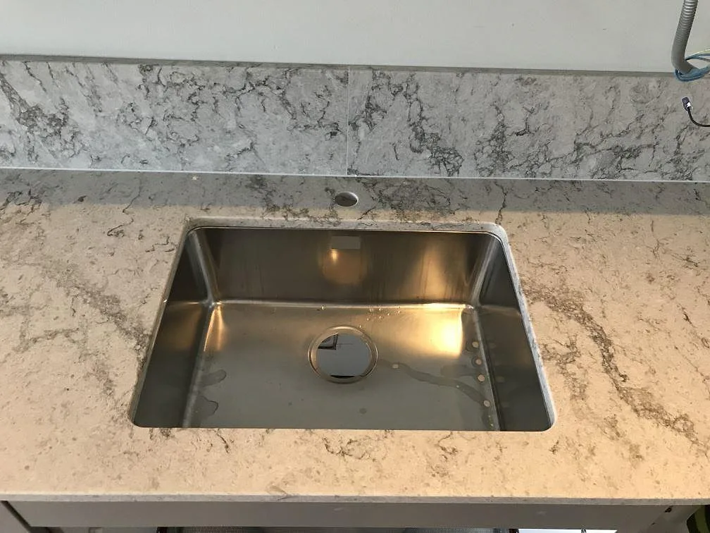 Underlimmad rostfri diskho i en köksbänkskiva av Caesarstone Moorland Fog med uppvikt stänkkant i polerad grå kvartskomposit i Västra Frölunda.