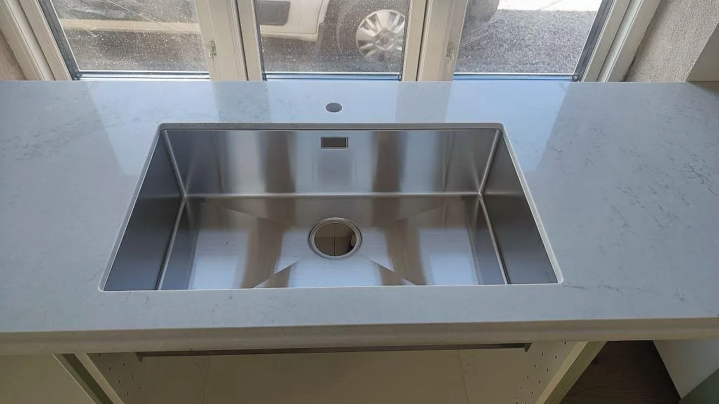 Underlimmad rostfri diskho monterad i polerad Caesarstone Noble Grey kompositbänkskiva vid fönster i Ekeby.