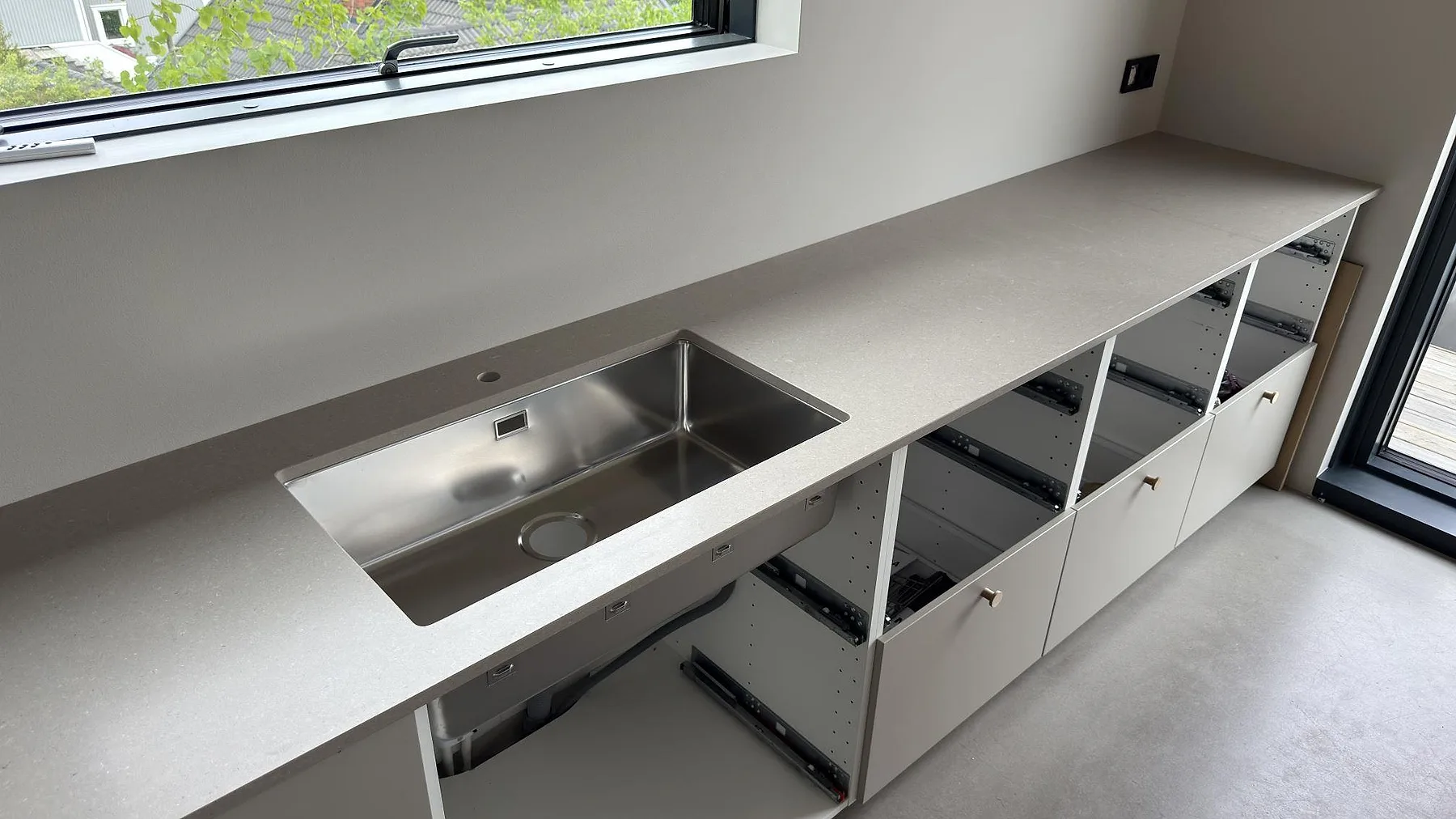 Köksbänkskiva i Caesarstone Shitake, en varm beige kvartskomposit med matt yta, monterad i en lång linje längs fönsterväggen med underlimmad rostfri diskho i ett kök under montering i Saltsjöboo.