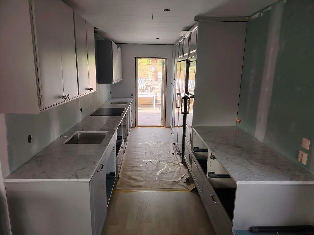 Köksbänkskivor i 20 mm polerad Carrara C-marmor längs två parallella bänkrader med synlig diskho, häll och stänkskydd i ett kök i Åkersberga.