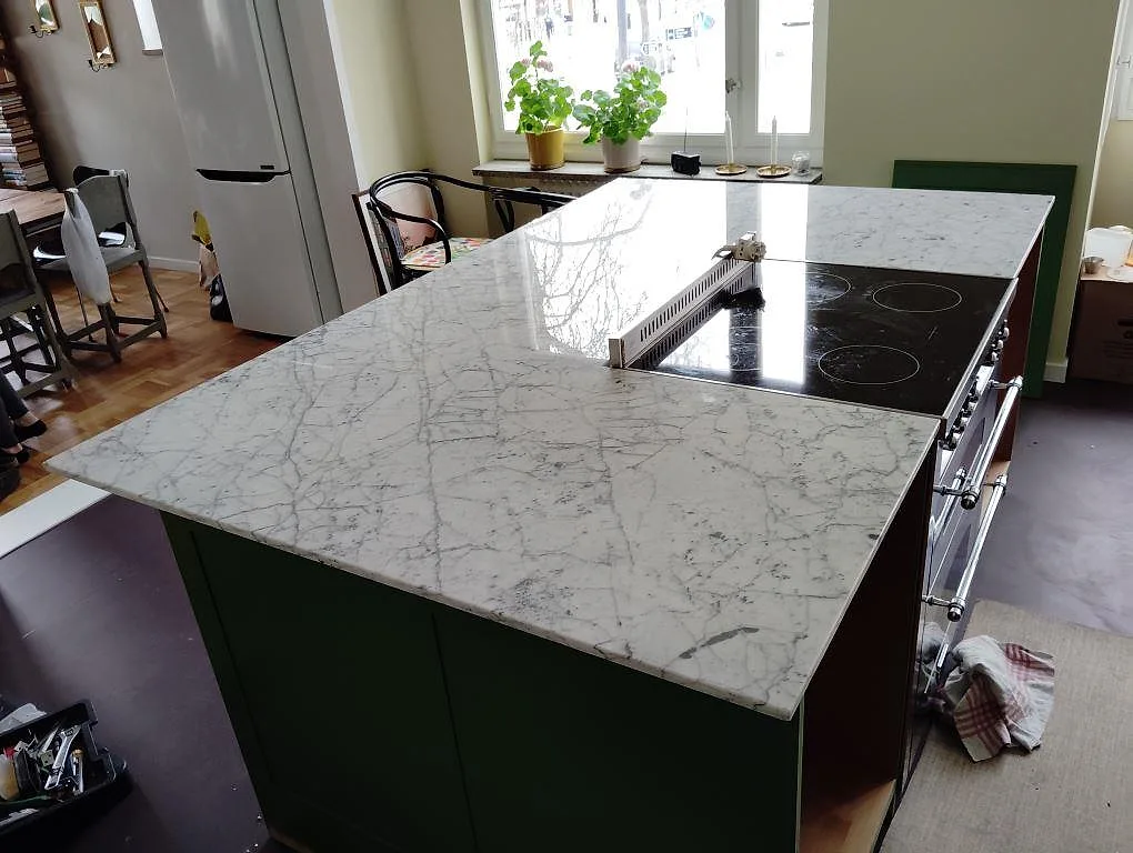Polerad köksbänkskiva i vit Carrara C marmor, 20 mm, på en stor köksö med svart häll och grön köksinredning i Årsta.