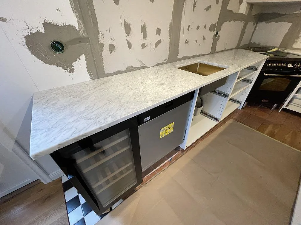 Matt köksbänkskiva i 30 mm Carrara C marmor längs väggen, med synligt urtag och dryckeskyl i ett kök i Älvsjö.