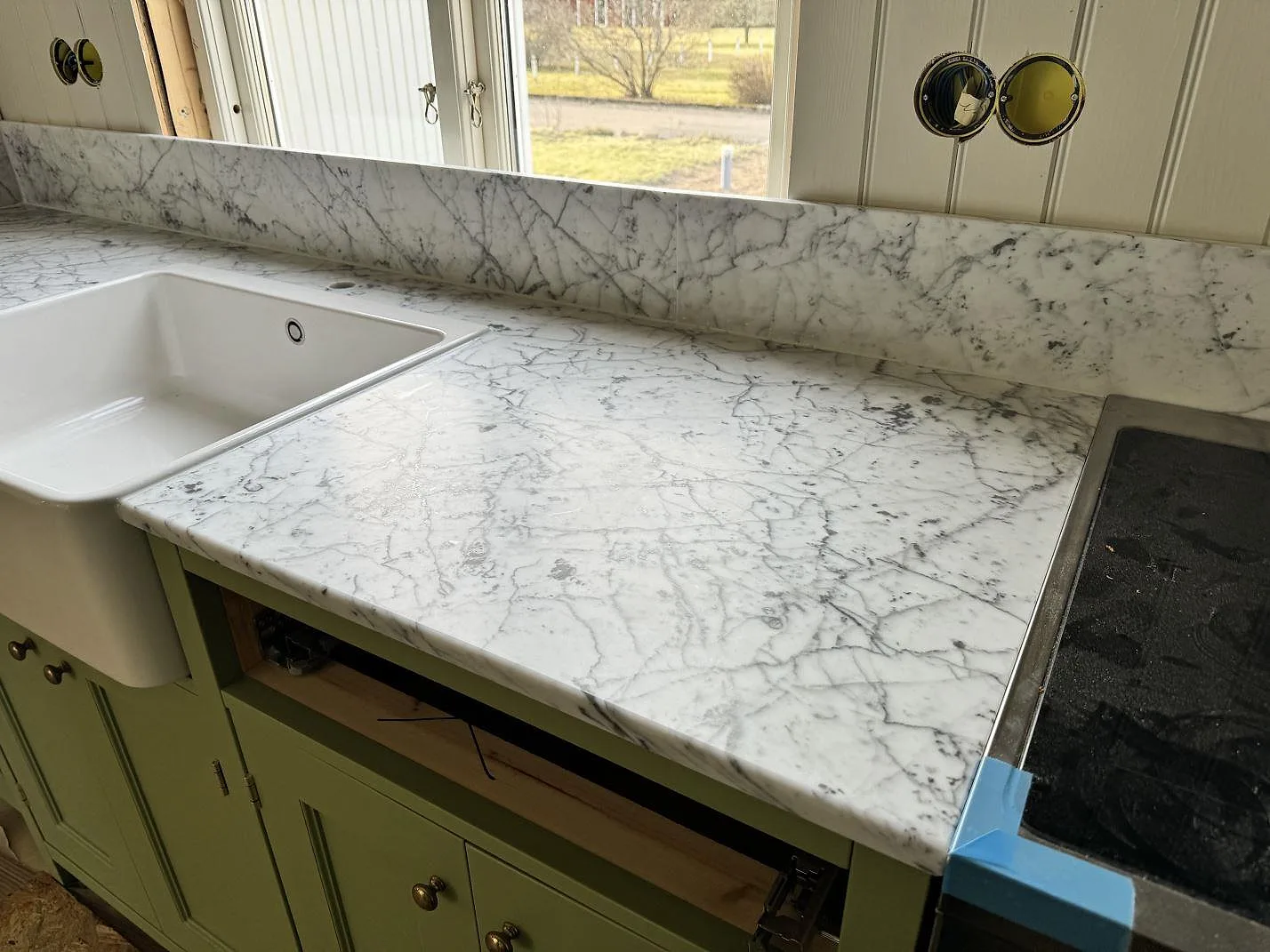 Köksbänkskiva i matt Carrara C marmor med vit keramisk diskho, infälld häll, låg stänkkant och sagegröna skåp i Alstermo.