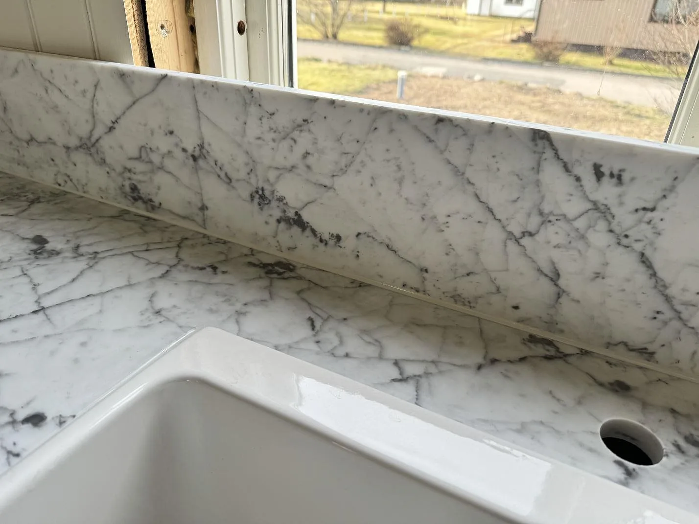 Närbild av vit keramisk diskho, blandarhål och uppvikt stänkkant i matt Carrara C marmor i köket i Alstermo.