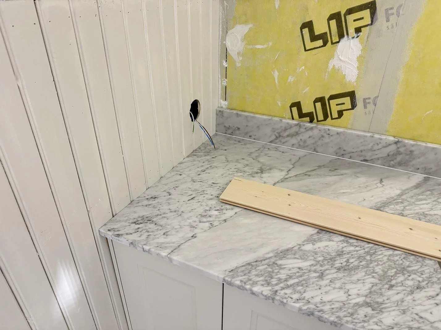 Marmorbänkskiva i Carrara C med låg uppvikt stänkkant mot vit pärlspontpanel, monterad över vita stommar i ett badrum under montering i Alstermo.