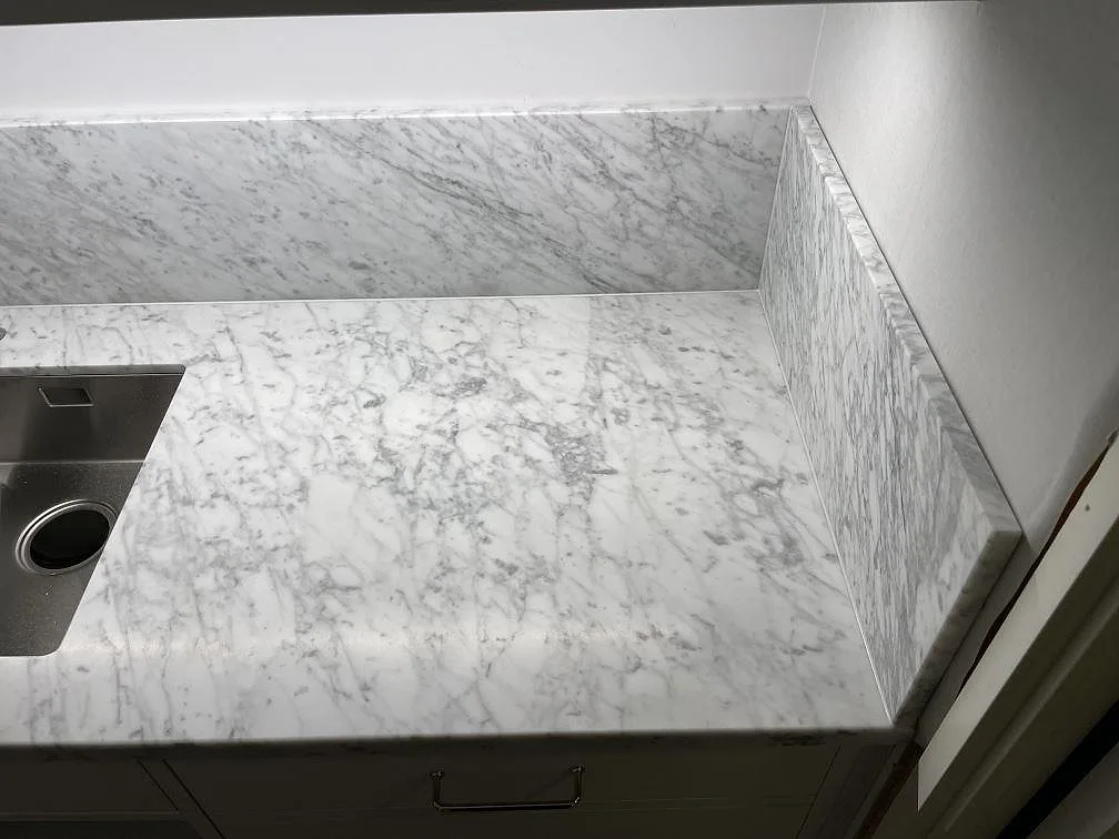 Detalj av matt Carrara C-marmor med bänkskiva, uppvikt stänkskydd och synlig ådring vid diskzonen i Avesta.