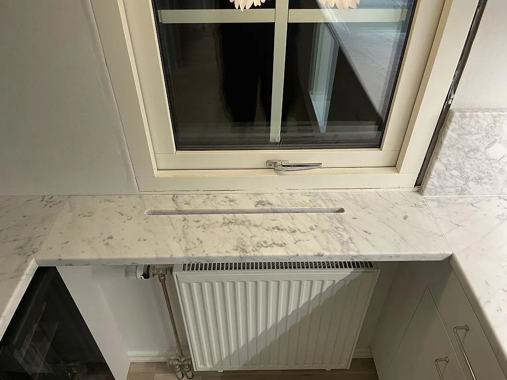 Fönsterbänk i matt Carrara C-marmor med ventilspår över radiator och anslutande köksbänk i Avesta.