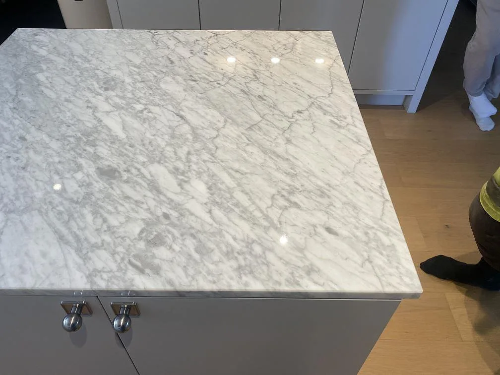 Detalj av köksö med polerad Carrara C marmorskiva i Bara, fotograferad under montering med kromknoppar på skåpen.