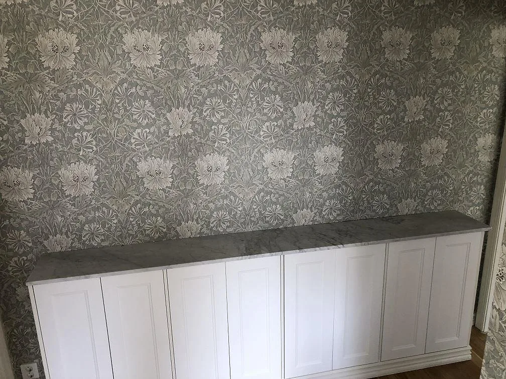 Bänkskiva i Carrara C, ljus marmor med grå ådring, monterad på en låg vit skåpsrad mot blommig tapet i Billdal.