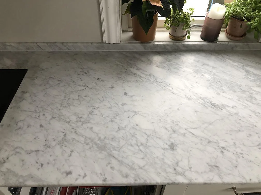Köksbänkskiva i matt vit Carrara C-marmor med grå ådring längs ett fönster med krukväxter i ett kök i Borås.