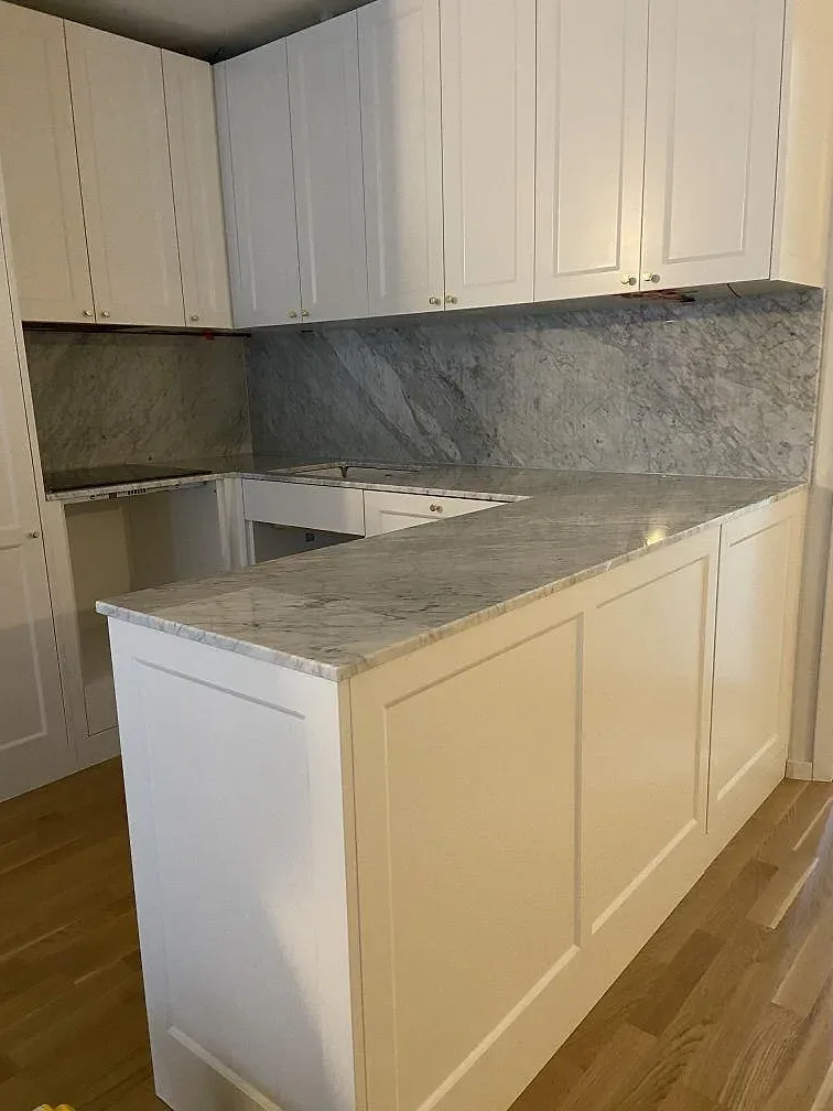 Polerad köksbänkskiva i Carrara C marmor med matchande väggbeklädnad och vita luckor i ett kök i Borås.