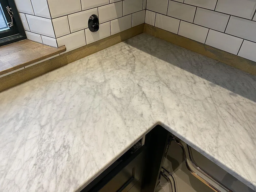Köksbänkskiva i matt Carrara C-marmor med grå ådring i L-format hörn mot vitt kakel i ett kök i Brantevik.