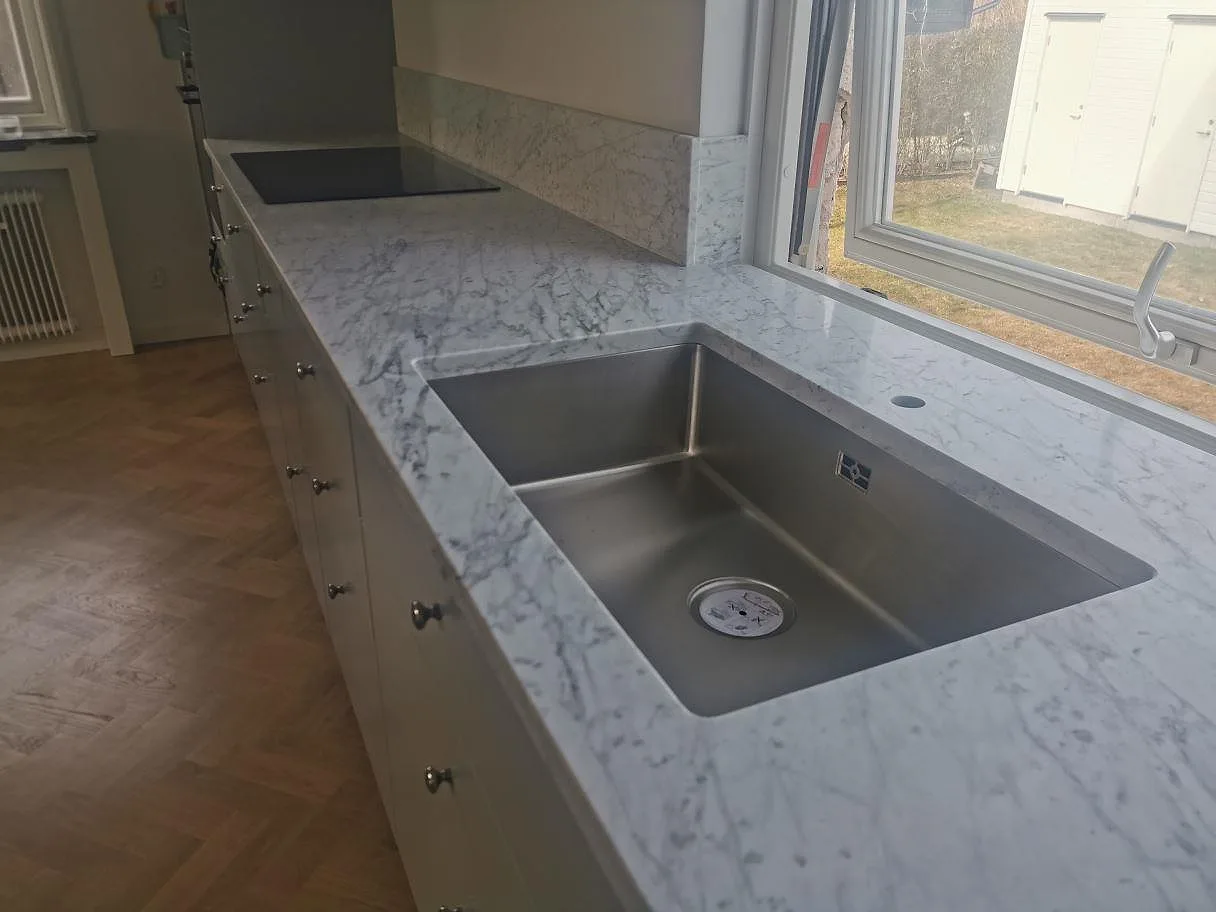 Köksbänkskiva i Carrara C marmor, matt yta, med undermonterad rostfri diskho och induktionshäll längs en lång bänkrad vid fönstret i ett kök i Bromma.