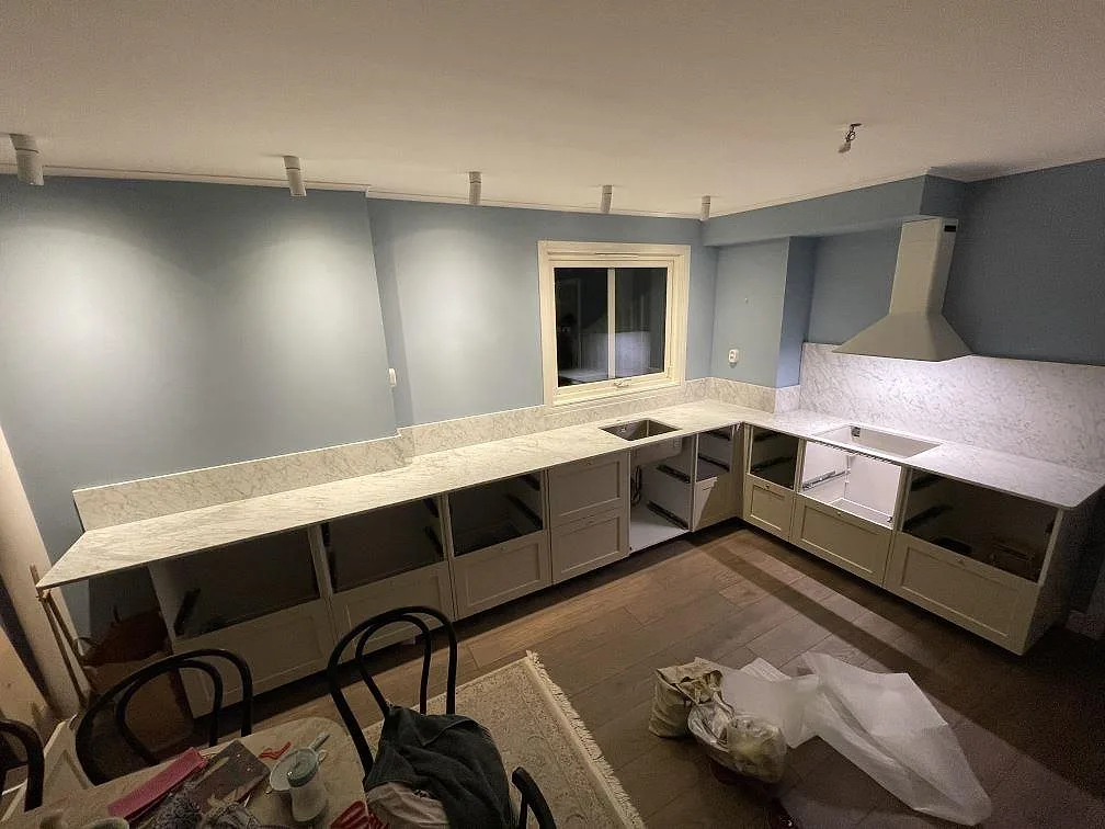 Köksbänkskiva i vit Carrara C marmor, matt, i ett L-format kök med duvblå väggar och gräddvita stommar i Bromma, fotograferat under montering.