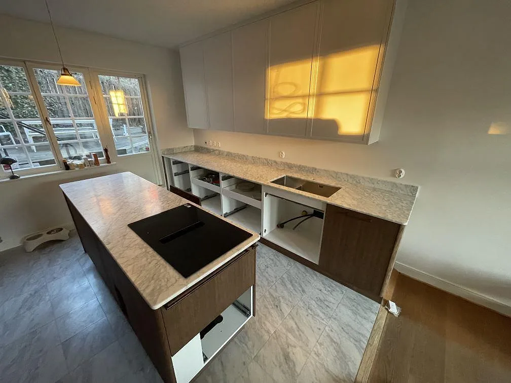 Köksbänkskiva i vit Carrara C marmor, matt yta, med underlimmad diskho längs väggen och en fristående köksö med induktionshäll i ett kök i Bromma.