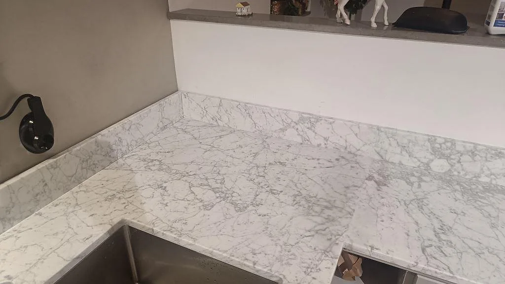 Hörndel av matt Carrara C marmorbänkskiva med diskho, stänkkant och tydlig ådring i köket i Bunkeflostrand.