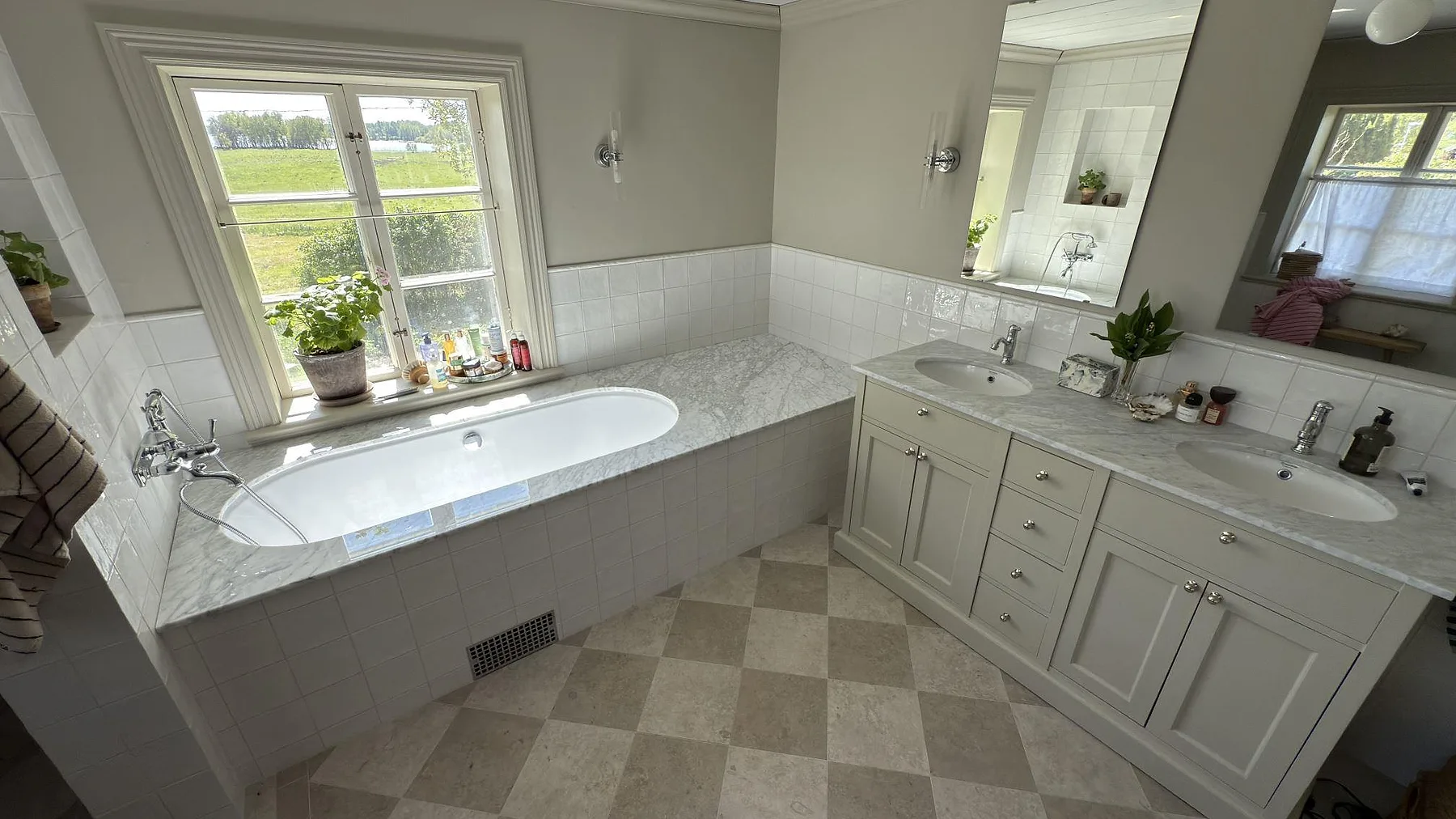 Helhetsbild av badrum i By Kyrkby med badkarssarg och kommod med dubbla tvättställ i Carrara C-marmor, kaklade väggar och schackrutigt stengolv.