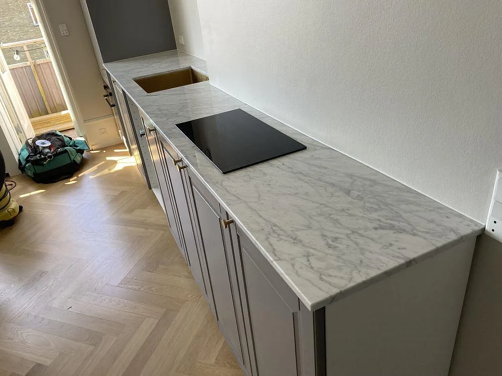 Köksbänkskiva i Carrara C marmor, 20 mm polerad, med svart häll och guldtonad diskho i ett kök under montering i Charlottenlund.