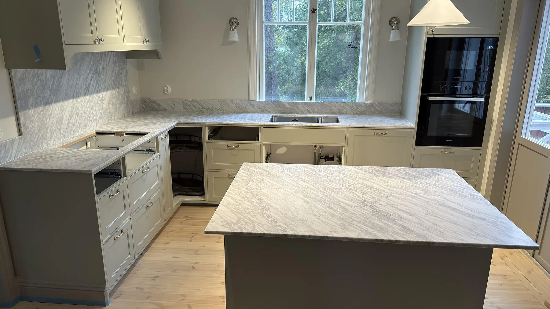 Matt köksbänkskiva och köksö i Carrara C marmor i ett kök i Djursholm, med stänkskydd, rostfri diskho och öppet hälluttag.