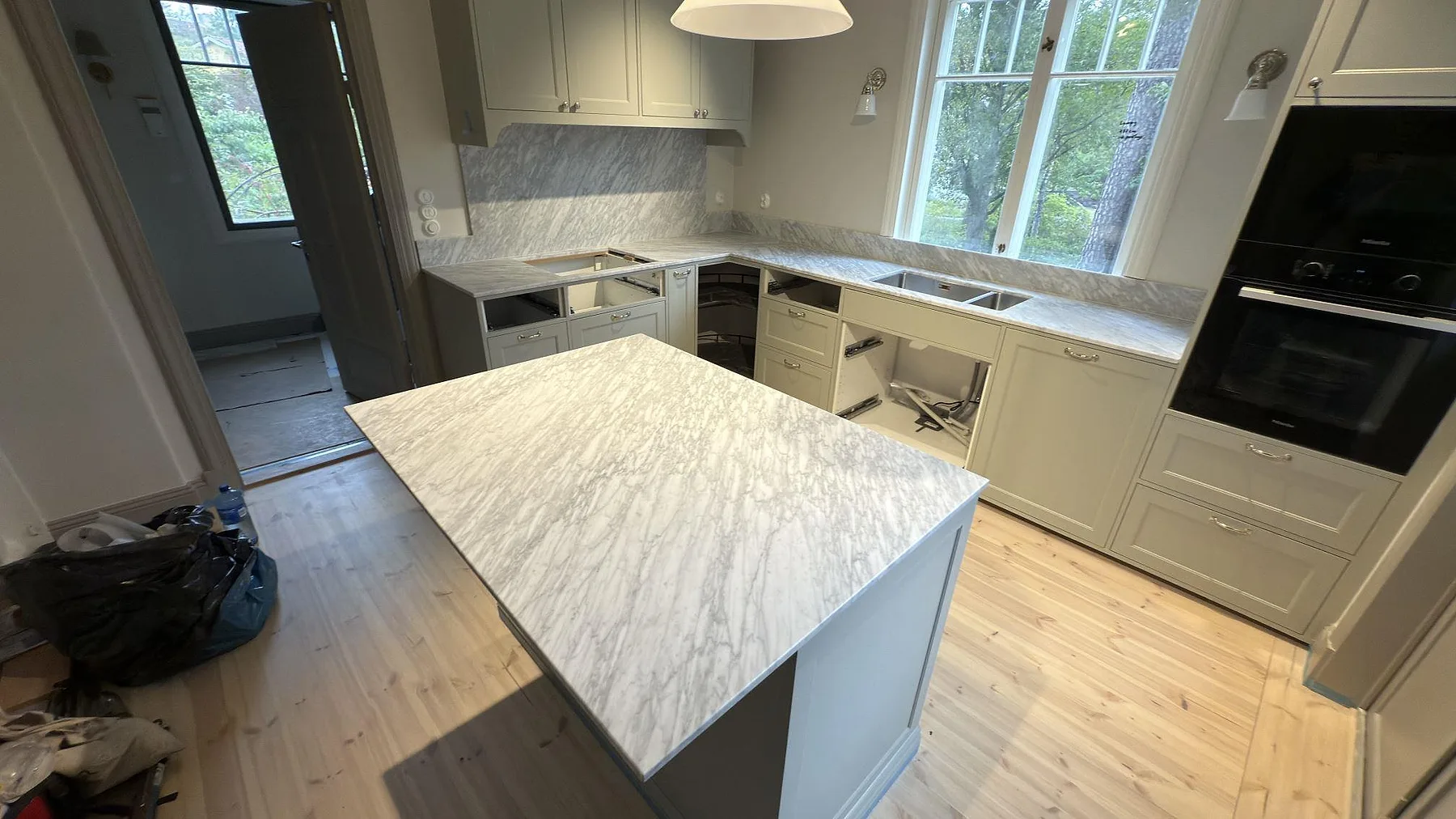 Översikt av matt köksö i Carrara C marmor i Djursholm, med väggbänkar, stänkskydd och hälluttag under montering.