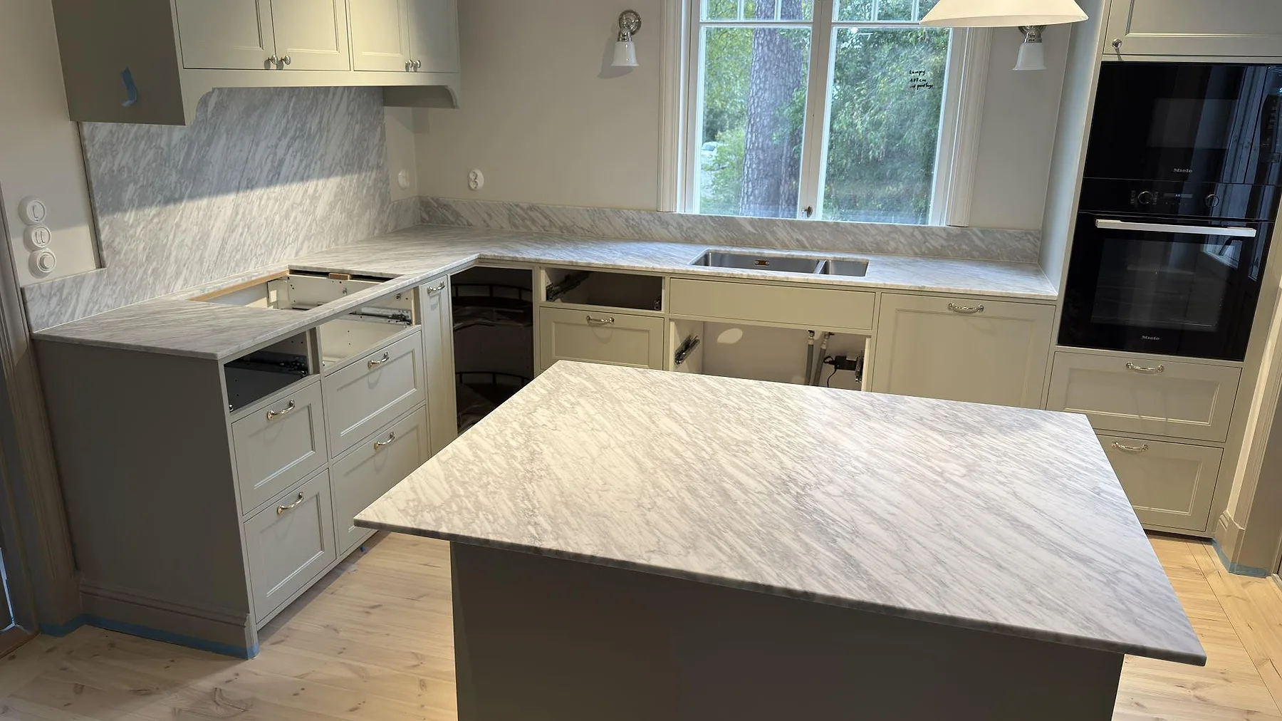Kök i Djursholm med matt Carrara C som marmor på bänkskivor, köksö och stänkskydd, med diskho vid fönstret.