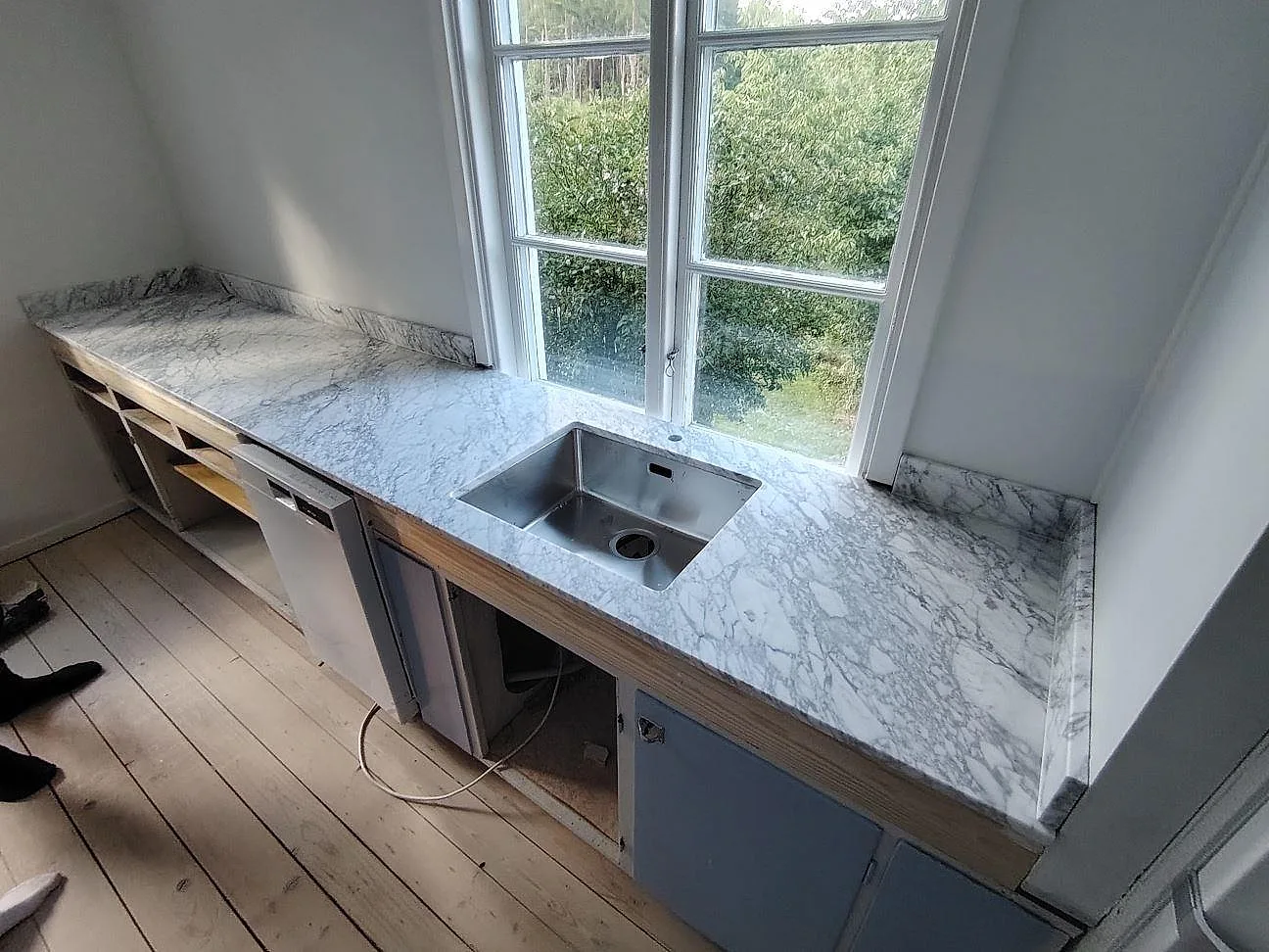 Köksbänkskiva i matt Carrara C-marmor, 20 mm, med rostfri diskho och uppvikt stänkkant under ett fönster i Edsbruk.