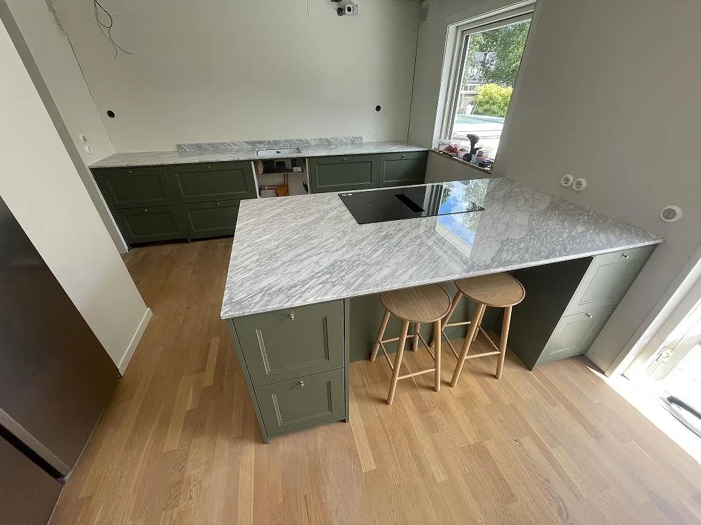 Köksbänkskiva i polerad Carrara C marmor med köksö, häll och väggbänk i ett kök i Ekerö.