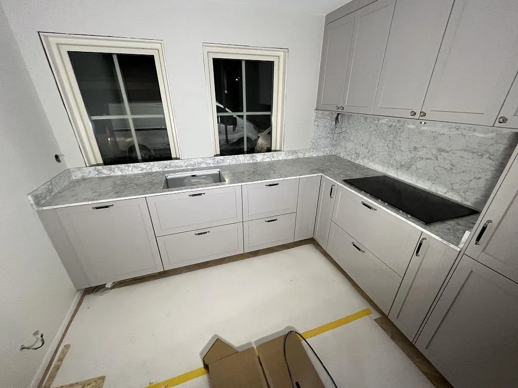 U-format kök med köksbänkskiva i matt Carrara C marmor, låg stänkkant, ho och svart häll i Enebyberg.