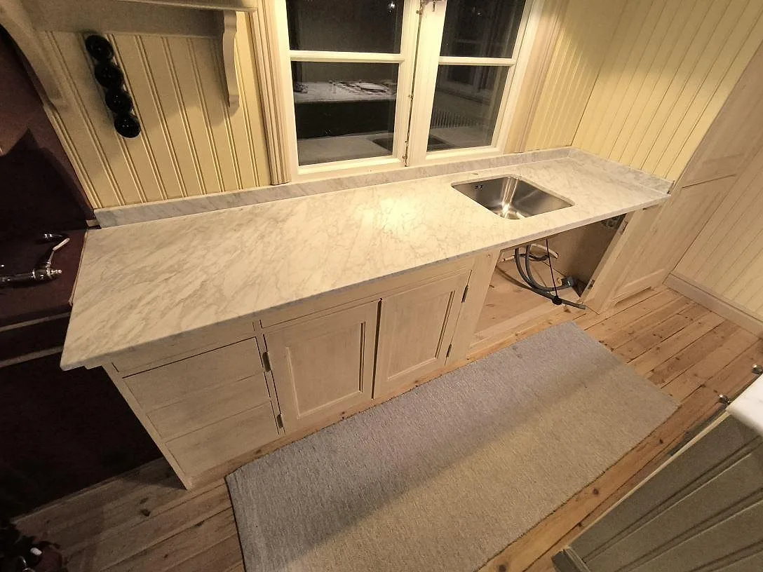 Köksbänkskiva i vit Carrara C marmor, matt, med låg stänkkant och rostfri diskho i ett kök i Enskede Gård.