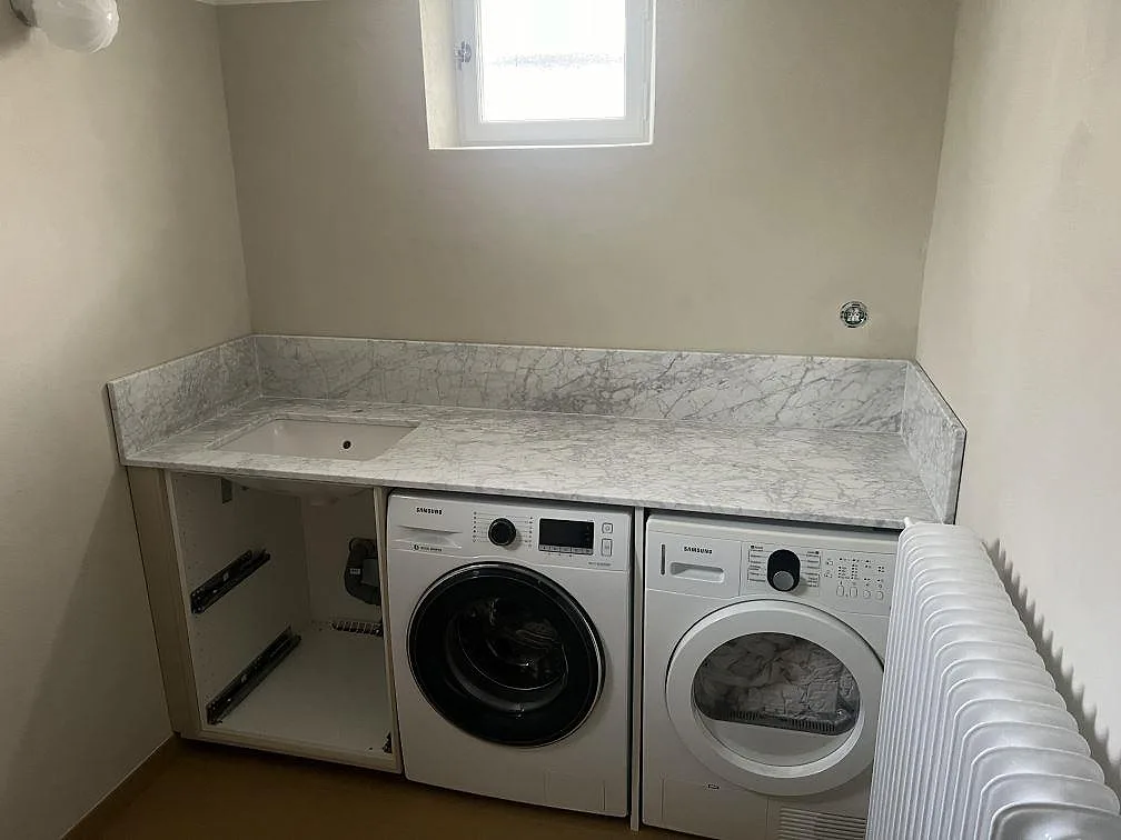 Bänkskiva i matt Carrara C marmor i en tvättstuga i Enskede, monterad över tvättmaskin och torktumlare med uppvikt kant.