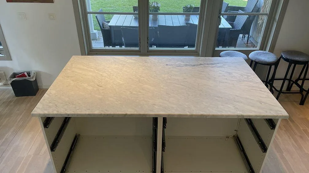 Köksö med bänkskiva i matt Carrara C marmor, grå ådring och öppna köksstommar under montering i Falsterbo.