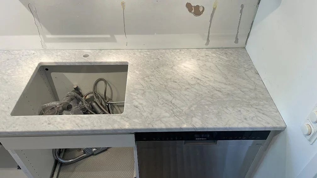 Höger del av väggbänk i matt Carrara C marmor med förberett urtag och diskmaskin under montering i Falsterbo.