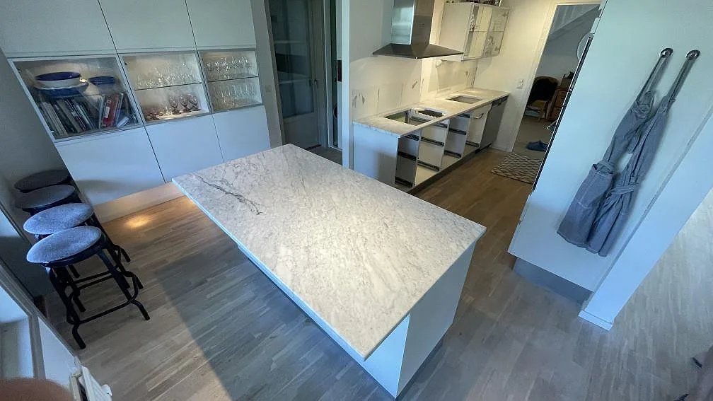 Köksbänkskiva i matt Carrara C marmor med bred köksö och väggbänk i ett ljust kök i Falsterbo.