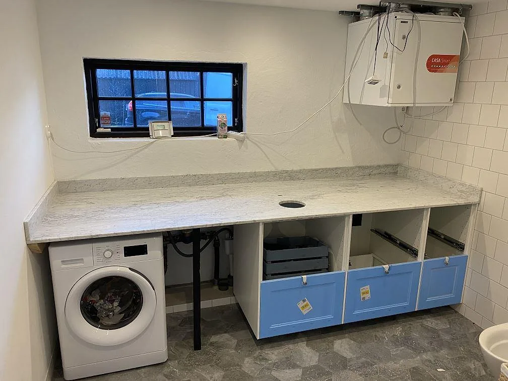 Marmorbänkskiva i matt Carrara C med låg stänkkant över tvättmaskin och badrumsstommar i Farhult.