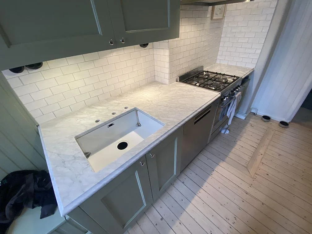 Köksbänkskiva i matt Carrara C marmor med underlimmad diskho, fristående spis och vitt kaklat stänkskydd i ett ljust kök i Göteborg.
